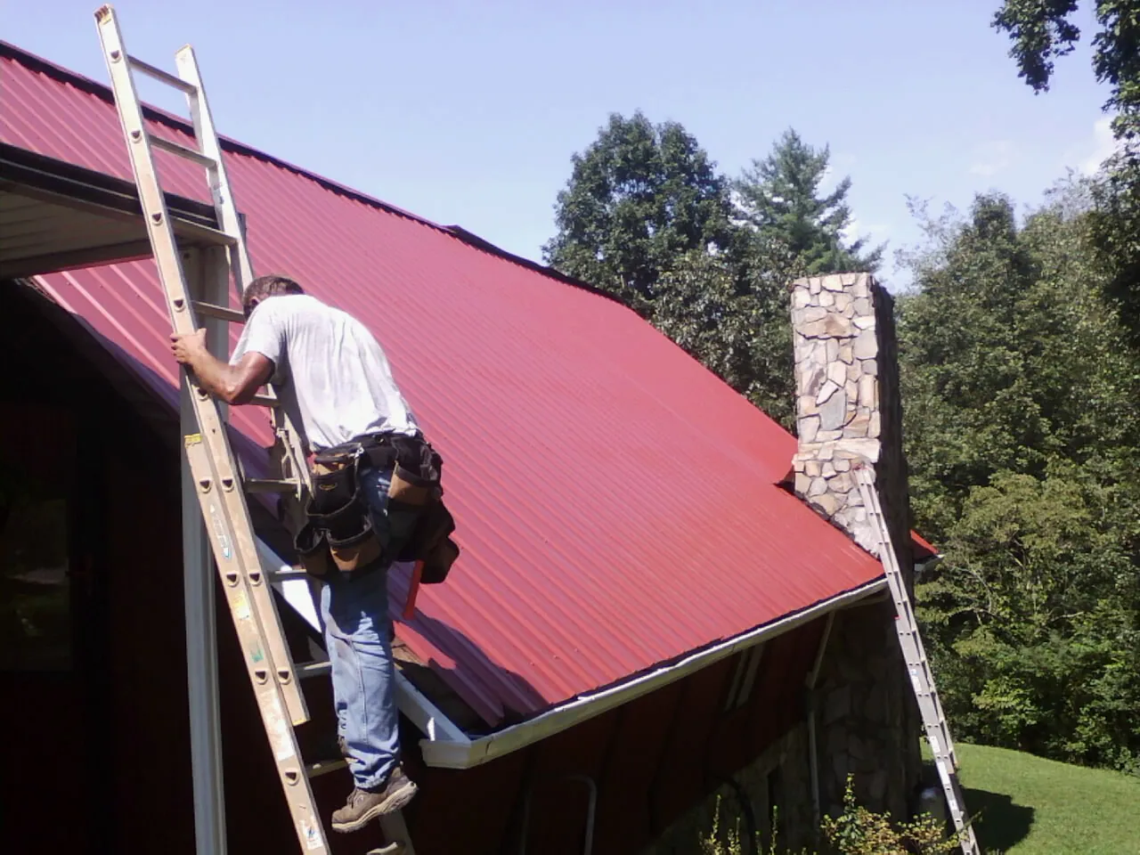 Roofer on a ladder beside a red metal roof and stone chimney — Soffit & Fascia Repair by Roofing Brothers in Nicollet